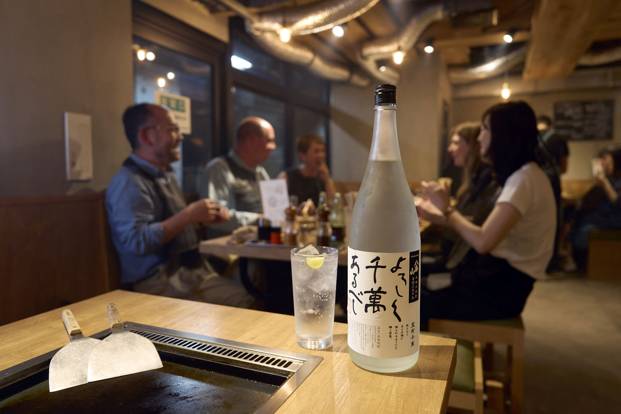 2F席の様子。米焼酎「よろしく千萬あるべし」のボトルと、「よろしく千萬すだ酎サワー」がテーブルの端に並ぶ。その奥には談笑するお客様。