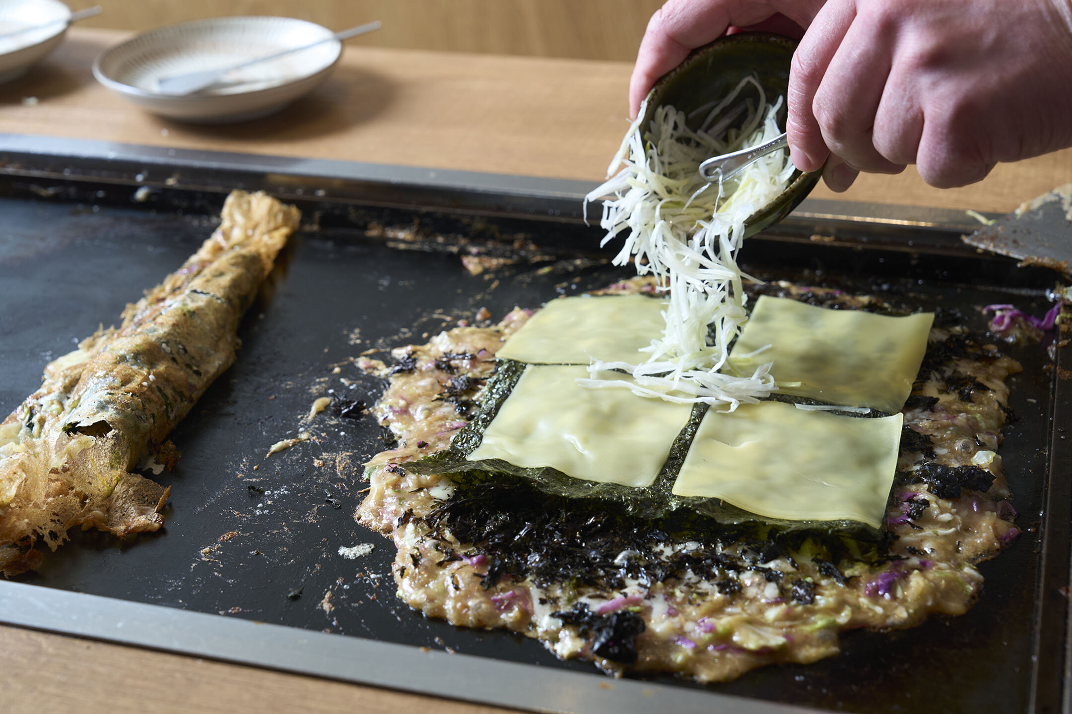 『バカノリチーズもんじゃ』。左がせんべいと呼ばれる棒状のもの。右はもんじゃの生地に海苔をのせ、スライスチーズ、さらにシュレッドチーズをかけている様子。