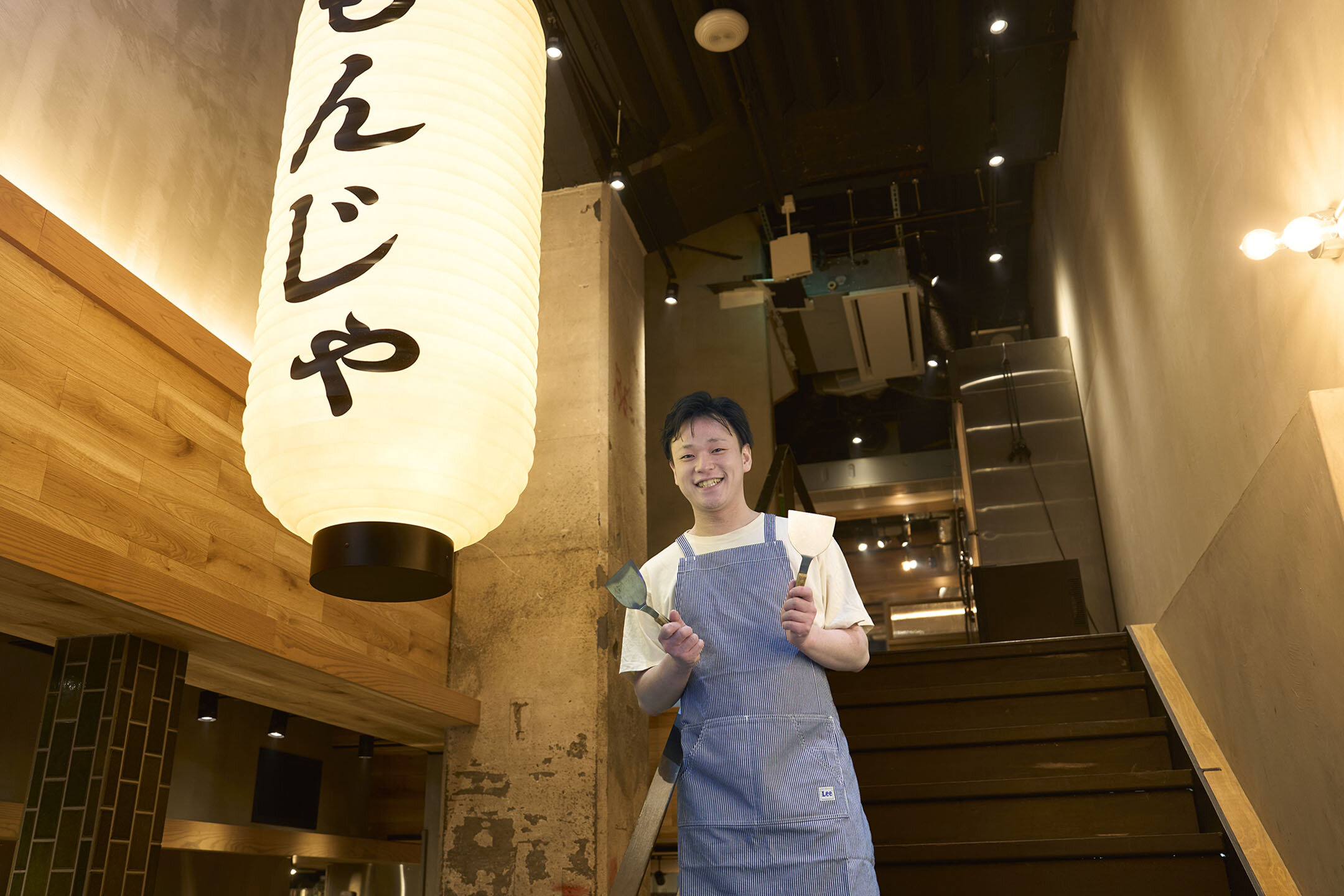 両手にへらを持ち、階段に立つ店長の曽根大吾さん。目の前には「もんじゃ」と書かれたちょうちんがぶら下がっている。