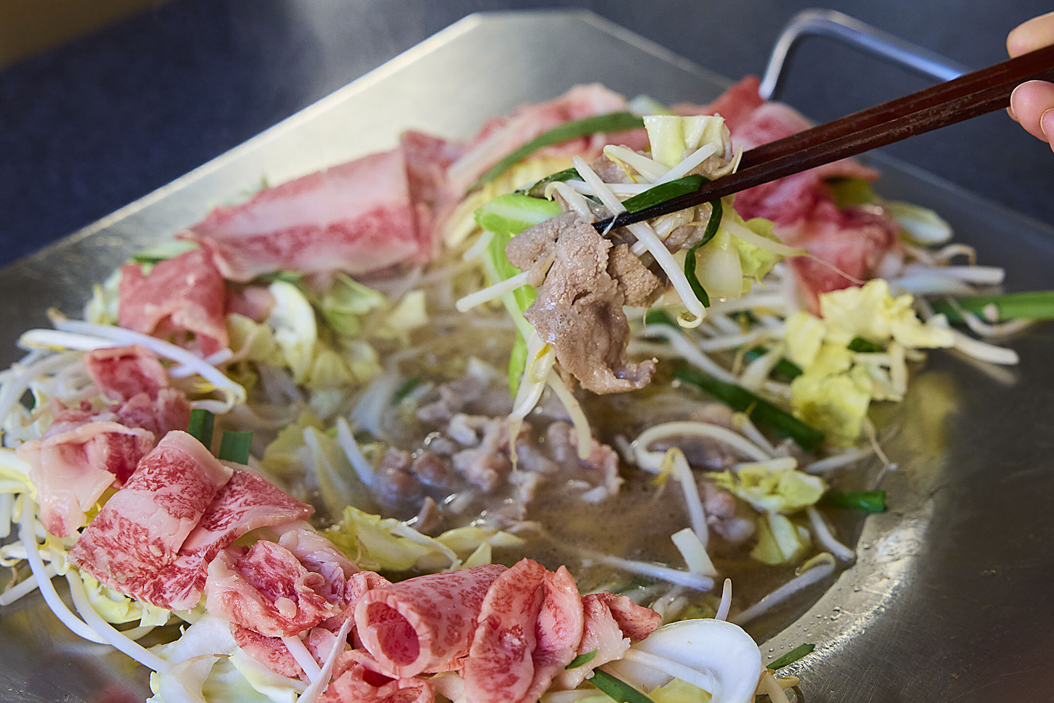 肉やもやしなどの野菜が出汁で煮込まれ、牛肉を箸で持ち上げている様子。